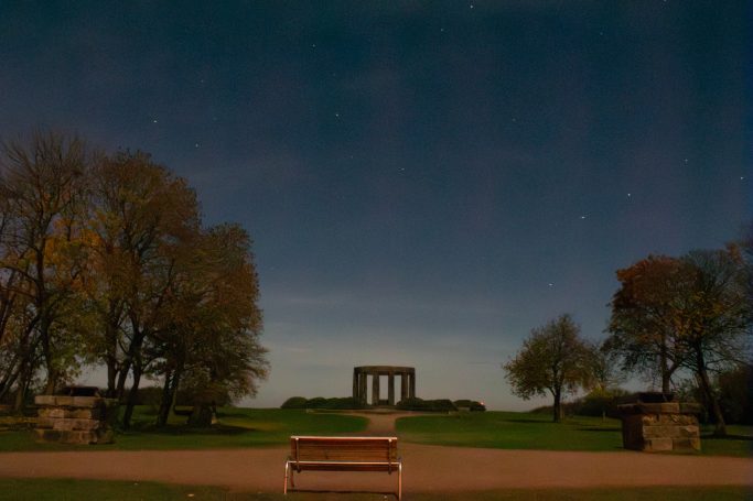 Blick auf einen Park mit einer Bank und einem monumentalen Bau im Hintergrund.
